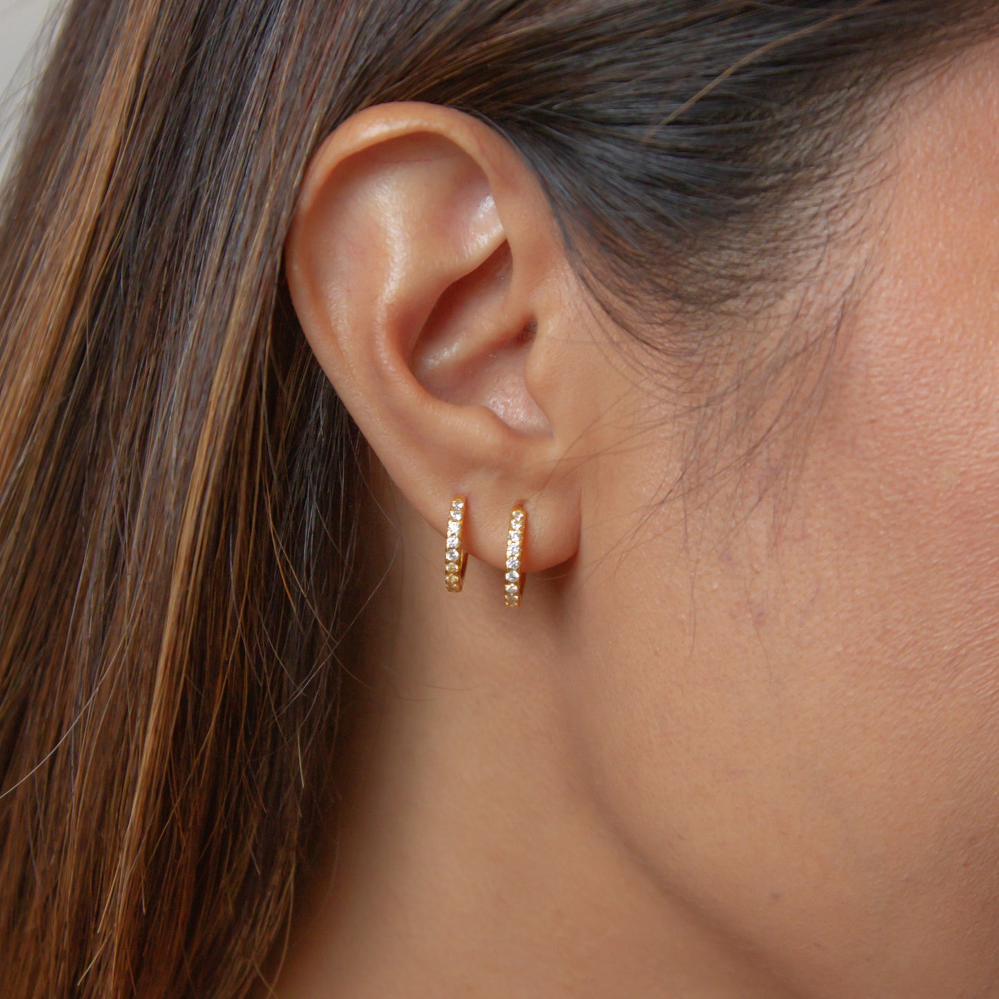photo of a woman's ear wearing two Soli huggies with white cubic zirconia stones made from sterling silver base, plated with 18k gold