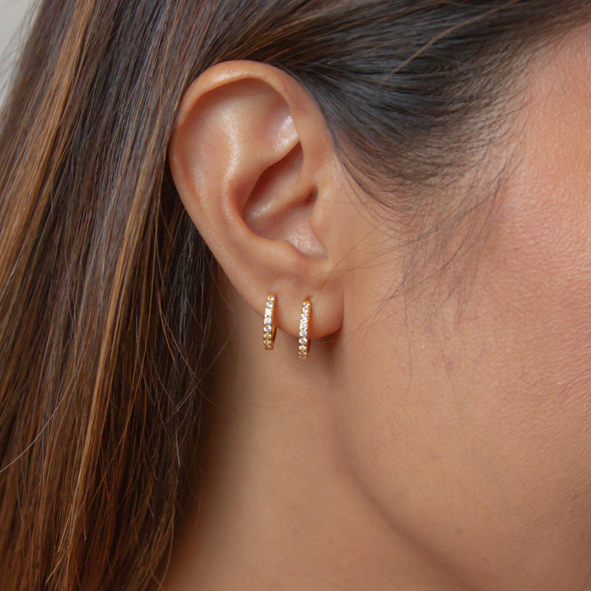 photo of a woman's ear wearing two Soli huggies with white cubic zirconia stones made from sterling silver base, plated with 18k gold